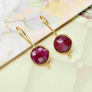 May include: A pair of gold-colored drop earrings with faceted, round, deep red gemstones. The earrings are set against a patterned background with floral and geometric designs. The gemstones have a brilliant cut.