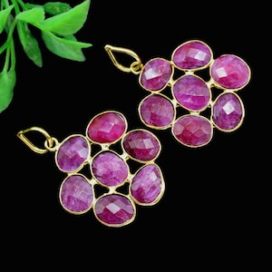 Könnte beinhalten: Zwei goldfarbene Anhänger, jeder mit einem Cluster aus ovalen, facettierten, tiefrosa Edelsteinen. Die Edelsteine sind in einer goldenen Lünette gefasst, und jeder Anhänger hat oben eine kleine Schlaufe zum Aufhängen. Der Hintergrund ist schwarz.