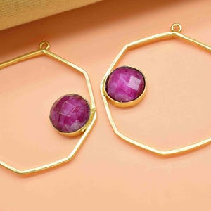 May include: Two gold-coloured, geometric-shaped earring components. Each earring features a faceted, round, magenta-coloured gemstone set within the gold frame. The earrings have a loop at the top for attachment.
