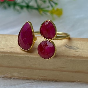 Può includere: Un anello dorato con tre pietre preziose rosse scuro sfaccettate. Una è a forma di goccia, le altre due sono rotonde. L'anello è appoggiato su una superficie di legno, con elementi floreali sfocati sullo sfondo.