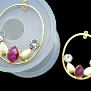 May include: Two gold-toned pendant earrings with an oval design. Each earring features two clear, round gemstones, two gold teardrop-shaped accents, and a central, faceted, ruby-colored gemstone. The earrings are displayed on a light gray surface against a black background.