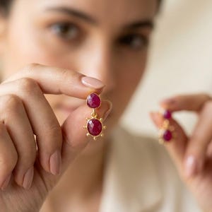 May include: A pair of gold-toned drop earrings featuring two faceted, deep pinkish-red ruby-like stones. The top stone is pear-shaped, while the bottom stone is oval-shaped and surrounded by a decorative sunburst-style metal frame with small beads.