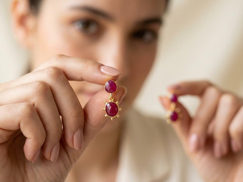 May include: A pair of gold-toned drop earrings featuring two faceted, deep pinkish-red ruby-like stones. The top stone is pear-shaped, while the bottom stone is oval-shaped and surrounded by a decorative sunburst-style metal frame with small beads.