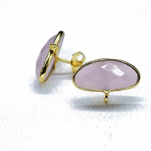 May include: A pair of pale pink gemstone stud earrings with a gold-coloured bezel setting. The earrings have a faceted surface and a post backing. The earrings are displayed on a white surface.