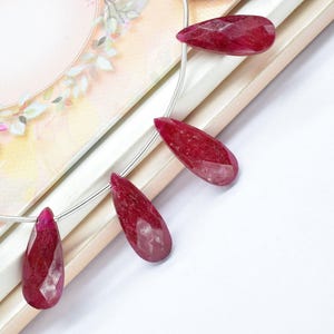 Puede incluir: Hilo de piedras preciosas de rubí facetadas en forma de lágrima. Las piedras de color rojo intenso están ensartadas en un fino alambre de plata, expuestas sobre un fondo decorado con motivos florales. Las piedras preciosas miden unos 3,8 cm de largo.