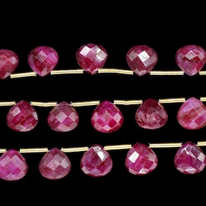 Pode incluir: Fios de contas de rubi vermelho escuro facetadas. As contas são enfiadas em fio dourado, com formato de gota nas fileiras inferiores e formato redondo na fileira superior. O fundo é preto.
