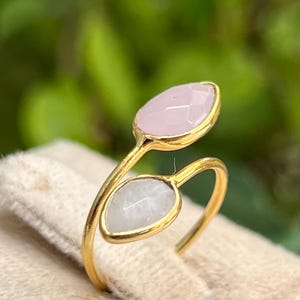 May include: A gold-colored ring featuring two teardrop-shaped gemstones, one pink and one white. The gemstones are set in gold bezels and arranged in a delicate, open design. The ring is displayed on a soft, beige surface, with a blurred green background.