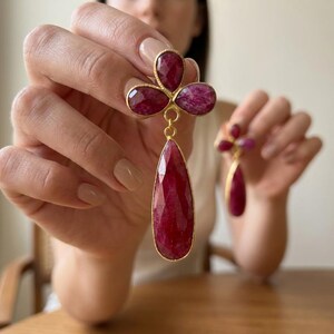 May include: A pair of gold-toned drop earrings featuring faceted, deep red ruby-colored stones. Each earring consists of a three-petaled clover-like top section connected to a single, elongated teardrop-shaped stone hanging below in a gold setting.