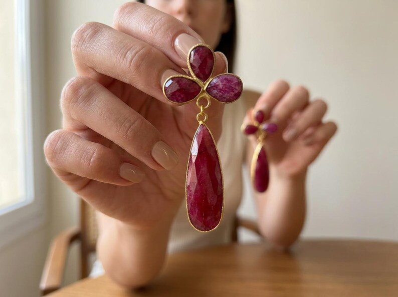 May include: A pair of gold-toned drop earrings featuring faceted, deep red ruby-colored stones. Each earring consists of a three-petaled clover-like top section connected to a single, elongated teardrop-shaped stone hanging below in a gold setting.