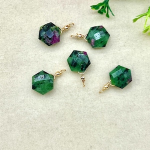 May include: Five faceted, hexagonal earrings with a green and red speckled pattern. Each earring is attached to a gold-coloured hook. The earrings are displayed on a cream-coloured surface, with pink and white flowers in the background.