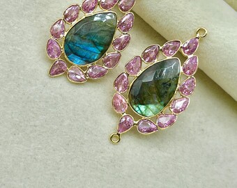 May include: Two gold-toned pendants, each featuring a teardrop-shaped labradorite stone with blue and green iridescence, surrounded by pink gemstones. The pendants have a small loop at the top for hanging.