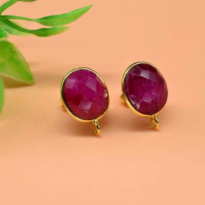 Pode incluir: Um par de brincos de pedra preciosa cor de rubi com engastes dourados. As pedras ovais têm uma superfície facetada. Os brincos são exibidos em um fundo cor de pêssego, com folhas verdes no canto superior esquerdo.
