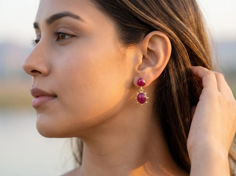 May include: A pair of gold drop earrings featuring two round, deep pink cabochon stones. The top stone is smaller and sits directly on the earlobe, while the larger bottom stone is suspended by a small gold link and framed by tiny gold spikes.
