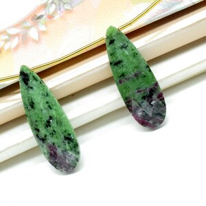 Op de afbeelding: Twee langwerpige, druppelvormige edelstenen. De stenen zijn groen met zwarte spikkels en een dieprode tint aan de basis. De edelstenen worden gepresenteerd op een wit oppervlak met een vleugje decoratieve achtergrond.