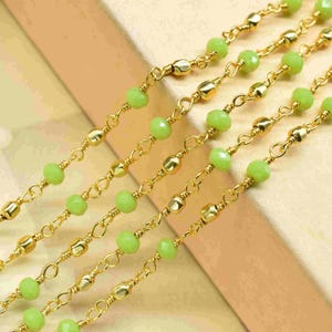 May include: Close-up of multiple strands of gold-colored chain necklaces. Each chain features small, round, light green beads interspersed with gold-colored, oblong beads. The chains are arranged diagonally across a light-colored surface.
