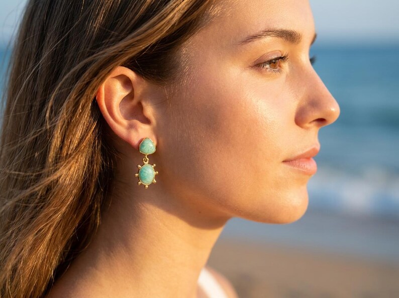 May include: A gold-toned drop earring featuring a small triangular turquoise-colored stone at the top and a larger oval turquoise-colored stone at the bottom, surrounded by a sunburst-style metal frame with small radiating spikes.