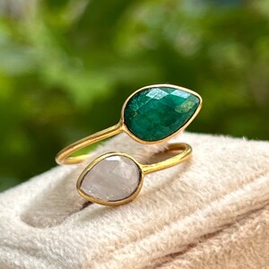 Pode incluir: Um anel dourado com duas pedras preciosas em forma de gota. Uma é verde esmeralda facetada, a outra uma pedra pálida e translúcida. O anel está sobre uma superfície macia e esbranquiçada.