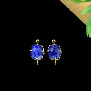 May include: Two faceted, square-shaped, deep blue gemstones with gold-colored metal settings. Each stone has a loop at the top and a small hook at the bottom. The background is black, with a hint of green and tan in the upper right corner.