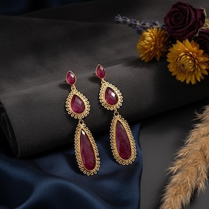 May include: A pair of gold-colored drop earrings with large, teardrop-shaped, ruby-red gemstones. The earrings have a filigree design around the gemstones and are displayed on a dark gray and navy blue background. A bouquet of flowers is in the background.