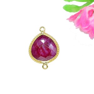 Op de afbeelding: Een druppelvormige, gefacetteerde robijn edelsteen hanger met een goudkleurige, touwtextuur rand. De hanger heeft twee kleine lussen om aan een ketting of andere sieraden te bevestigen. Een roze bloem met groene bladeren op de achtergrond.