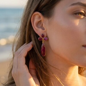 May include: A gold-toned earring featuring three small, faceted, deep pinkish-red stones arranged in a clover-like cluster at the top, with a single, larger, teardrop-shaped, faceted stone of the same color hanging vertically below.