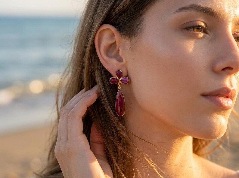 May include: A gold-toned earring featuring three small, faceted, deep pinkish-red stones arranged in a clover-like cluster at the top, with a single, larger, teardrop-shaped, faceted stone of the same color hanging vertically below.