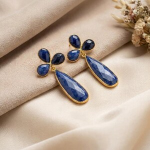 May include: A pair of gold-toned drop earrings featuring faceted, deep blue teardrop-shaped stones suspended from a three-petaled floral cluster of smaller, matching blue stones. The metal settings have a bright, polished gold finish.