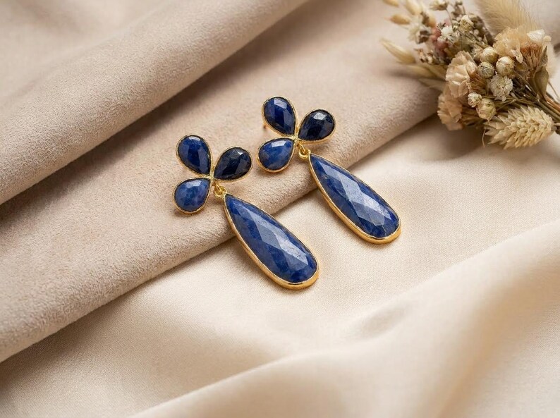 May include: A pair of gold-toned drop earrings featuring faceted, deep blue teardrop-shaped stones suspended from a three-petaled floral cluster of smaller, matching blue stones. The metal settings have a bright, polished gold finish.