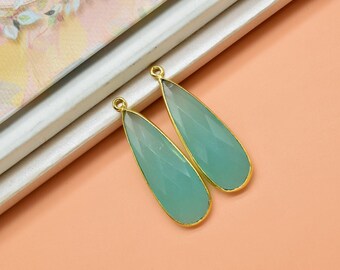 May include: Two teardrop-shaped, faceted, aqua-colored gemstones with gold-colored metal edging. The gemstones are displayed on a peach-colored surface, near a white and floral-patterned object. The gemstones have a loop at the top for hanging.