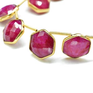 Puede incluir: Primer plano de un hilo de piedras preciosas de rubí facetadas y hexagonales engastadas en biseles de oro. Los rubíes son de un color rojo-rosa intenso y vibrante, y los engastes de oro proporcionan un cálido contraste. Las piedras preciosas están ensartadas en un fino hilo de oro.