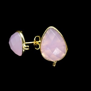 May include: A pair of stud earrings featuring light pink, teardrop-shaped gemstones set in gold-coloured metal frames. The earrings have a post and butterfly backing. The background is black.