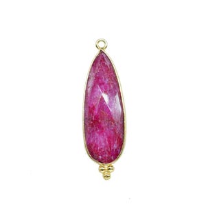 Peut inclure: Un pendentif en forme de goutte d'eau avec une pierre précieuse rose foncé à facettes, sertie dans un cadre en métal doré. Le pendentif a une petite boucle en haut et trois petites sphères décoratives en bas.