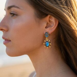 May include: A gold-toned drop earring featuring two bezel-set labradorite stones. The top stone is teardrop-shaped, while the bottom stone is oval with a sunburst-inspired metal frame featuring small spheres around the outer edge.