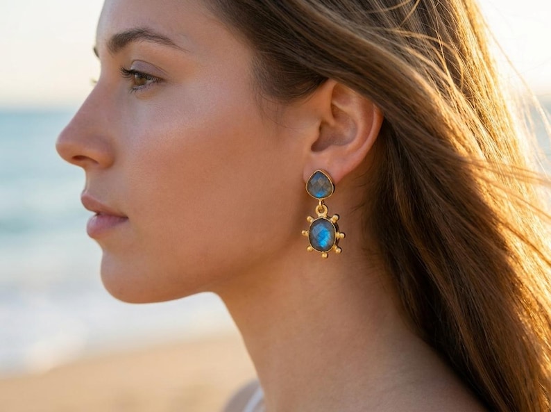 May include: A gold-toned drop earring featuring two bezel-set labradorite stones. The top stone is teardrop-shaped, while the bottom stone is oval with a sunburst-inspired metal frame featuring small spheres around the outer edge.