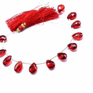 May include: A strand of faceted, teardrop-shaped red gemstones. The gems are strung on a thin, silver-colored wire, with a red tassel at the top. The beads are a deep, translucent red color, and the tassel is a vibrant, solid red.