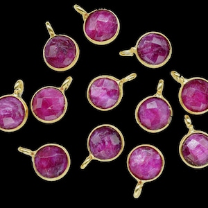 Può includere: Una collezione di pendenti con pietre preziose di rubino con montature color oro. Ogni pendente rotondo presenta un rubino rosa scuro sfaccettato. I pendenti hanno un piccolo anello nella parte superiore per il fissaggio a una catena o ad altri componenti di gioielleria. Lo sfondo è nero.