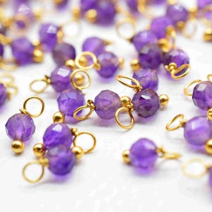 Puede incluir: Primer plano de cuentas de piedras preciosas de amatista con detalles dorados. Las cuentas moradas facetadas están ensartadas con pequeños bucles dorados. Las cuentas están dispersas sobre una superficie blanca.