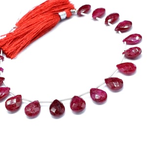 May include: A strand of faceted, teardrop-shaped ruby gemstones. The deep red stones are strung on a thin, silver-colored wire. A red tassel is attached to the end of the strand. The gemstones are displayed against a white background.