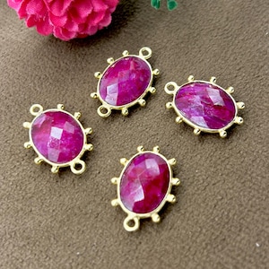 Könnte beinhalten: Vier ovale, goldfarbene Anhänger mit facettierten, tiefrosa Edelsteinen. Jeder Anhänger hat eine Schlaufe oben und unten zur Befestigung und einen dekorativen Rand. Sie liegen auf einer braunen Oberfläche.