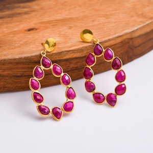 May include: A pair of gold-colored teardrop earrings. Each earring features a gold stud and a teardrop-shaped frame filled with vibrant, magenta-colored gemstones. The earrings are displayed on a wooden surface.