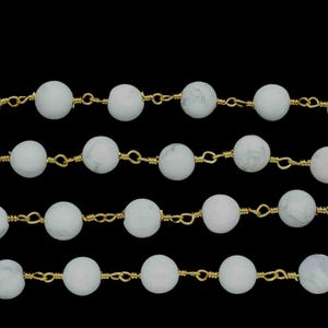 May include: Close-up of multiple strands of white, round beads strung on gold-colored wire. The beads have a matte finish and are evenly spaced. The background is solid black.