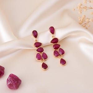 Può includere: Orecchini dorati con gemme rosse a forma di goccia. Gli orecchini sono esposti su un tessuto di raso color crema, con due pietre di rubino grezze e fiori secchi sullo sfondo. Un elegante gioiello.