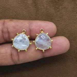 Peut inclure: Deux pendentifs triangulaires dorés avec un centre en pierre claire et facettée. Chaque pendentif a une petite boucle en haut pour l'accrocher et est entouré de petites perles dorées. Les pendentifs sont présentés sur un fond marron.