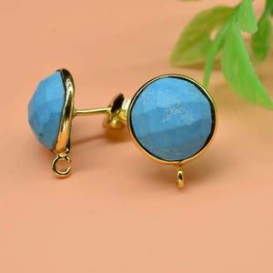 May include: A pair of gold-colored stud earrings with round, light blue, faceted stones. The earrings have a post and loop design. The background is a light peach color with green leaves.