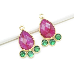 May include: Two gold-toned teardrop-shaped pendants. Each pendant features a large, faceted, magenta-colored gemstone at the top, with three smaller, round, green gemstones below. The pendants are set against a white background.