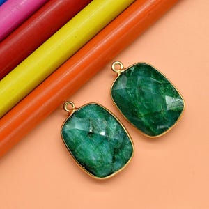 May include: Two emerald green gemstone pendants with gold-colored frames. The pendants are rectangular with rounded corners and a faceted surface. The background includes colorful pencils in blue, pink, red, yellow, and orange.