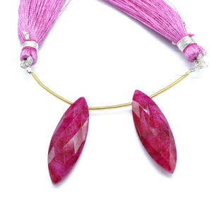May include: Two faceted, elongated ruby-coloured gemstone beads are suspended from a gold-coloured wire, with a pink tassel at each end. The beads have a pointed, leaf-like shape. The background is white.