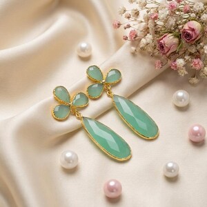 May include: A pair of dangle earrings featuring light green, faceted, teardrop-shaped stones suspended from three-petaled, clover-like green stone studs. Each component is framed in a thin gold-toned metal setting, creating a cohesive design.