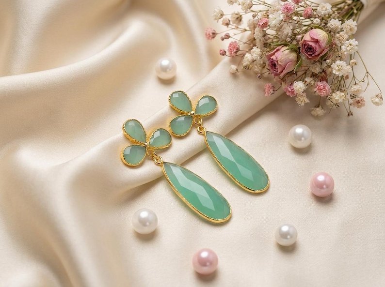 May include: A pair of dangle earrings featuring light green, faceted, teardrop-shaped stones suspended from three-petaled, clover-like green stone studs. Each component is framed in a thin gold-toned metal setting, creating a cohesive design.
