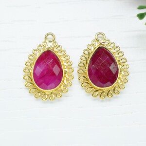 Puede incluir: Dos colgantes dorados en forma de lágrima con un centro de piedra preciosa rosa intenso facetada. Los colgantes tienen un borde calado decorativo. El fondo es una superficie blanca con algunas flores amarillas.